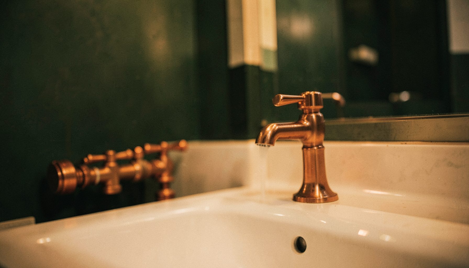 A warm, softly lit residential bathroom with copper plumbing fixtures and a deep green accent wall